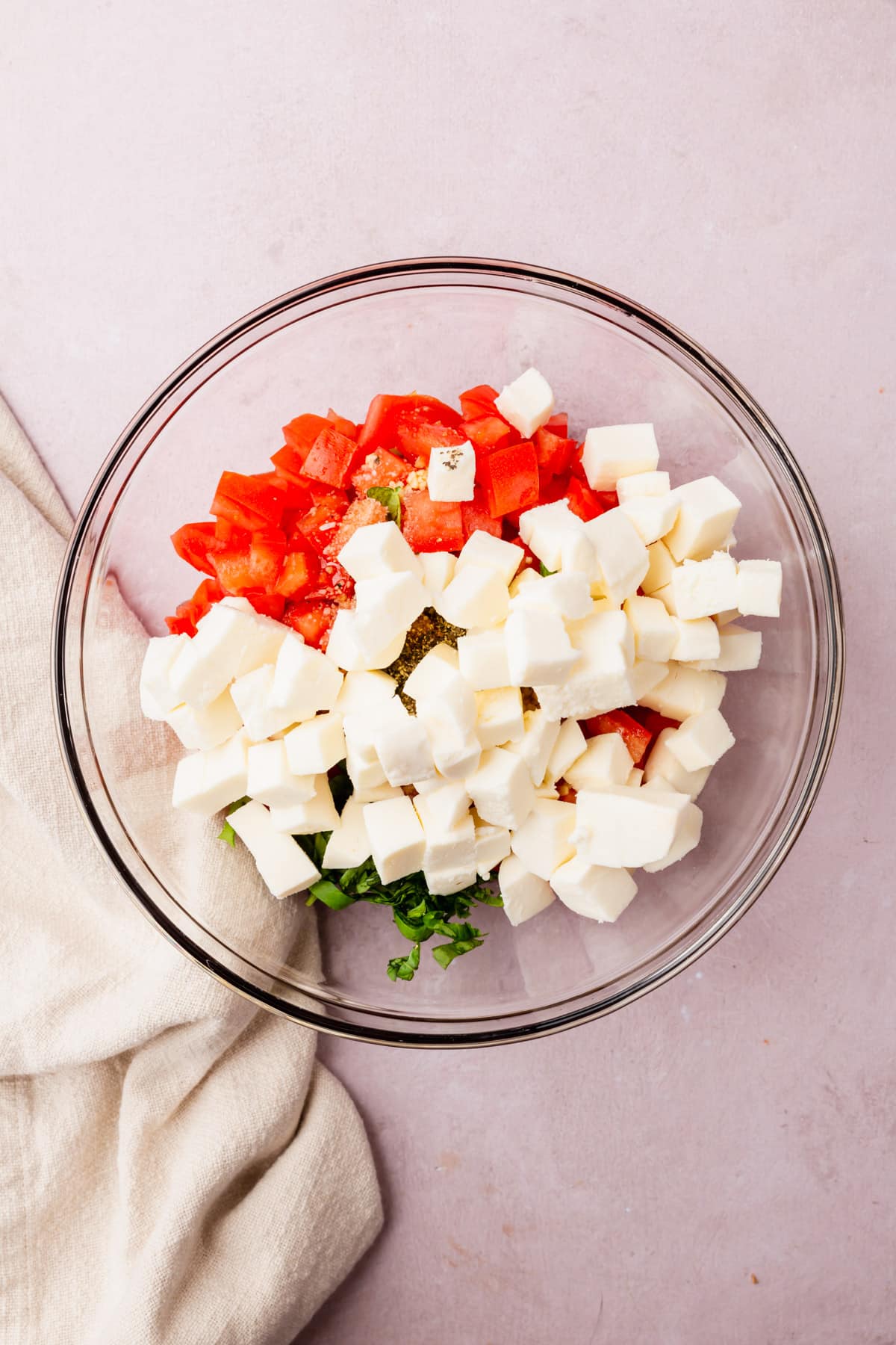 A glass mixing bowl with diced tomatoes, sliced basil, diced fresh mozzarella and minced garlic before mixing together.