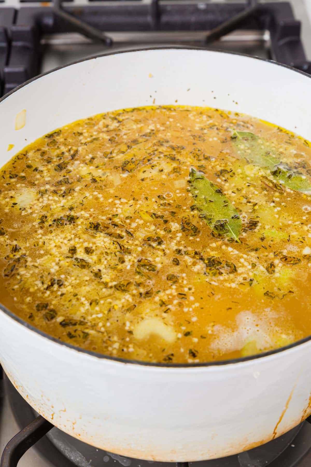 A white dutch oven of chicken broth, spices, garlic, and bay leaves simmering on the stove top.