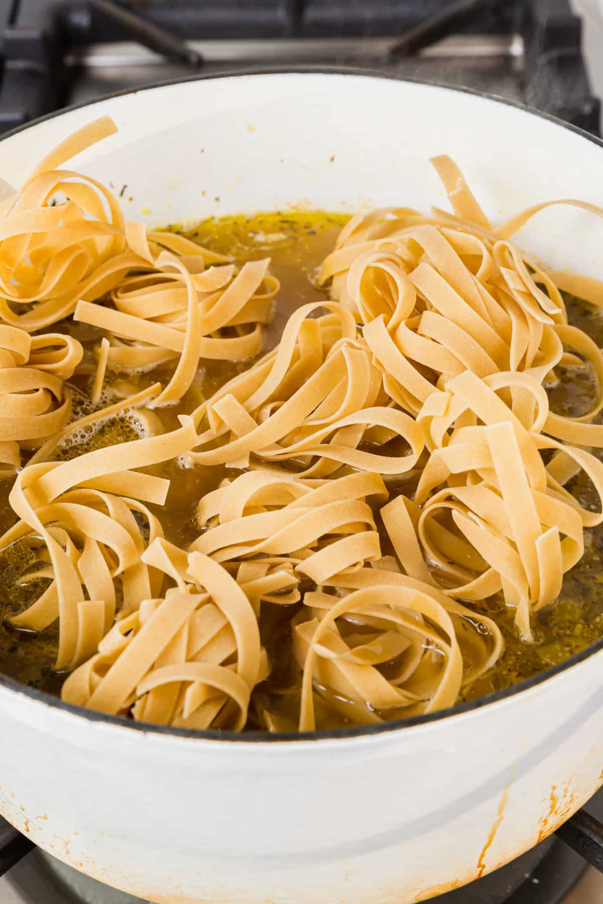 A white dutch oven of gluten free egg noodles added to a pot of chicken noodle soup before the noodles have been cooked.