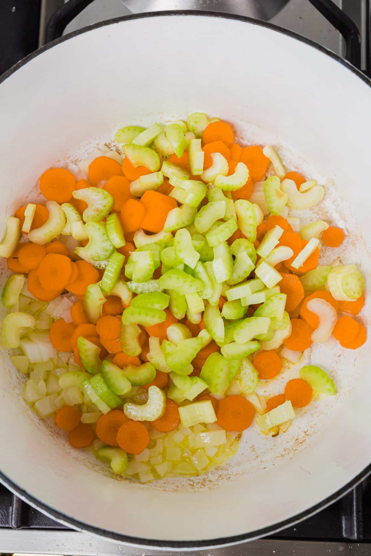An overhead view of a white dutch oven with raw diced celery, carrots and onion in olive oil before cooking.