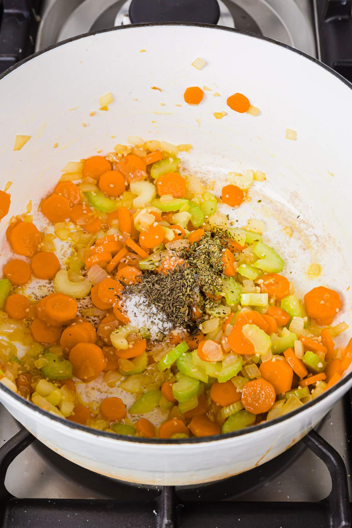 A white dutch oven with sliced carrots, celery, diced onion, salt, dried herbs and minced garlic cooked in olive oil.