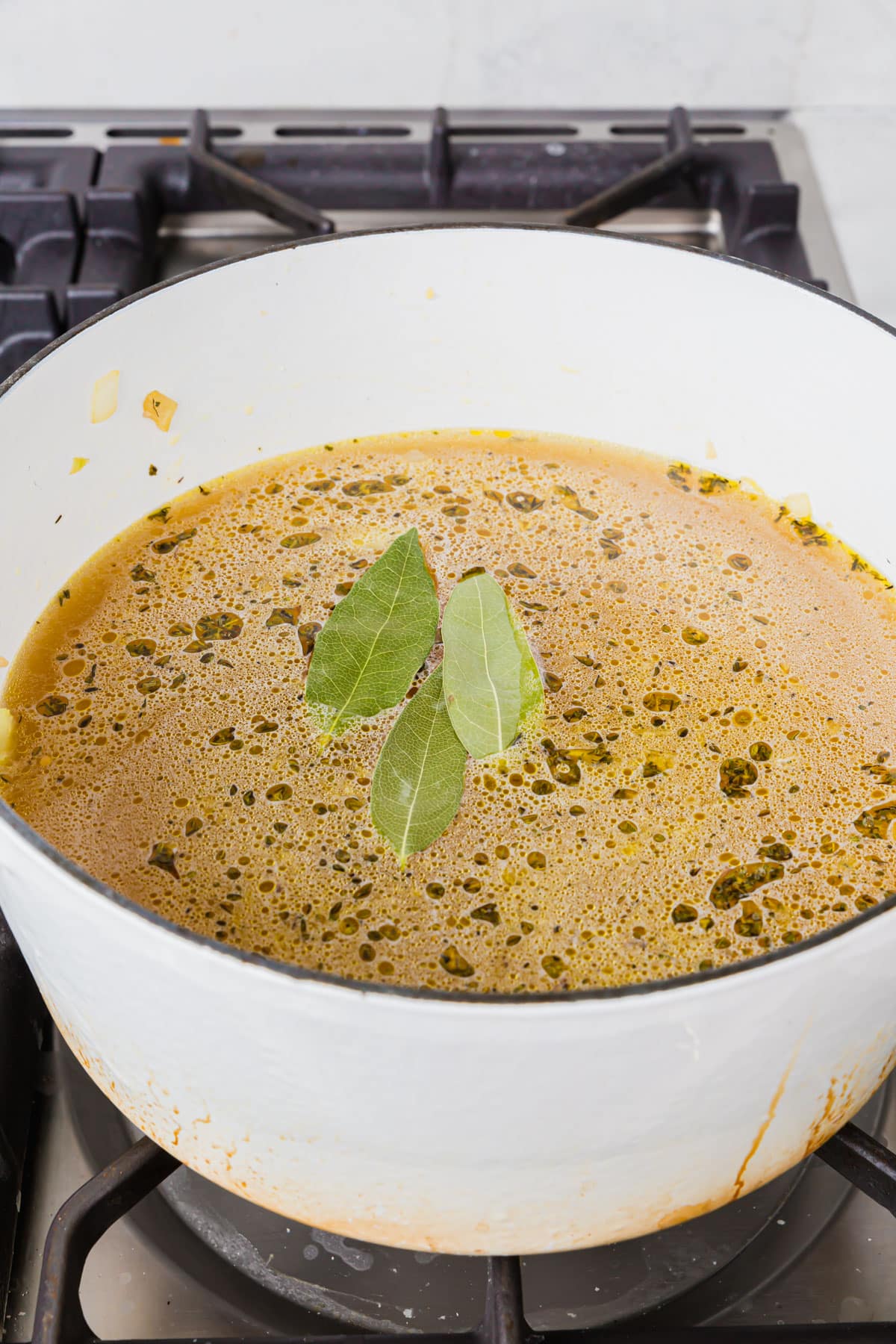 A white dutch oven of chicken broth, herbs, diced onion and dried bay leaves.