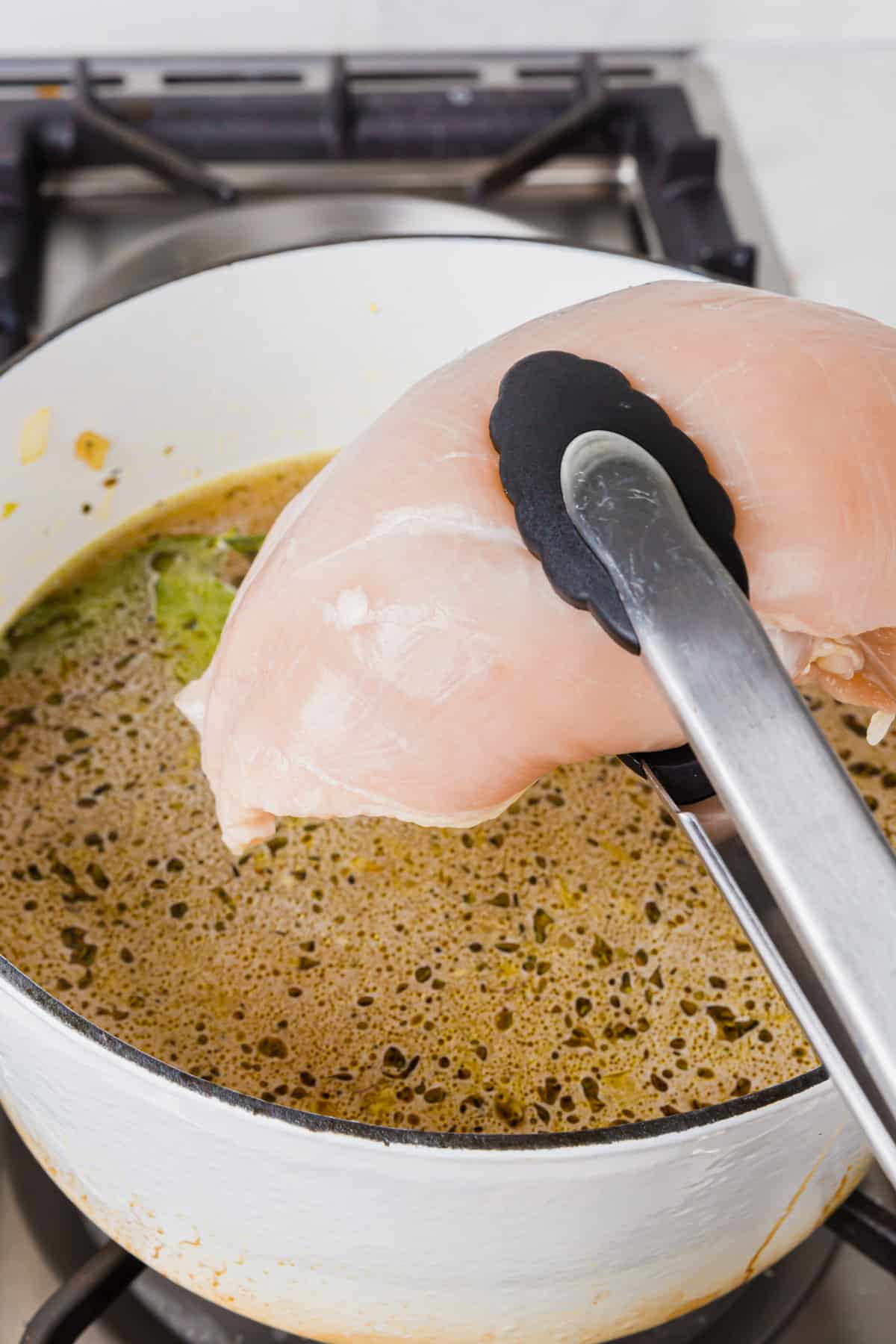 A pair of tongs lowering a raw chicken breast into a dutch oven of chicken broth, herbs, bay leaves, and minced garlic.