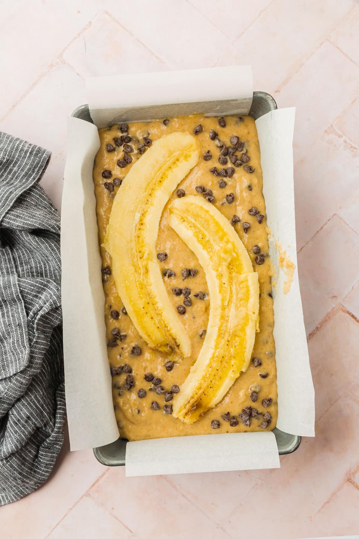 A loaf pan with gluten free banana bread batter with mini chocolate chips and two cross sections of a banana on top before baking in the oven.