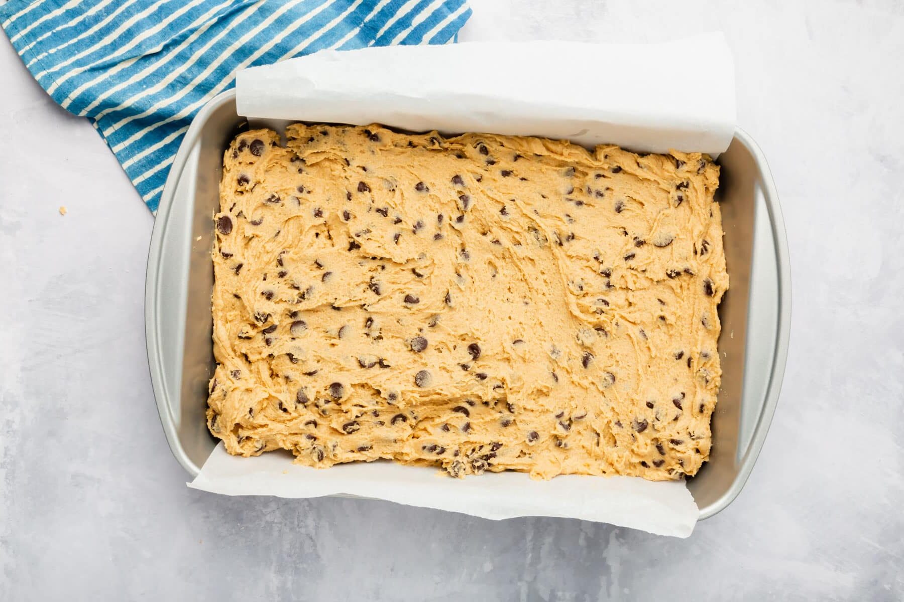 A rectangle metal baking pan lined with parchment paper and filled with gluten free chocolate chip cookie bar dough before baking in the oven.