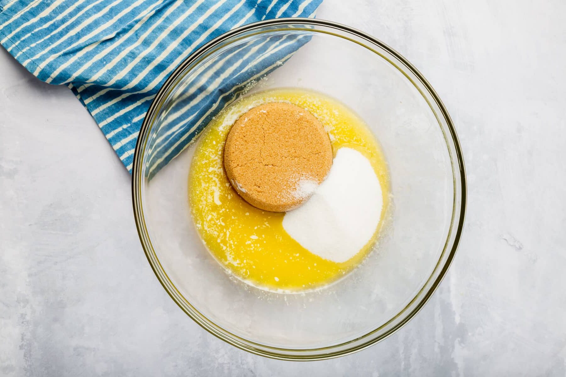 A glass mixing bowl with melted butter topped with granulated sugar and brown sugar before mixing together.