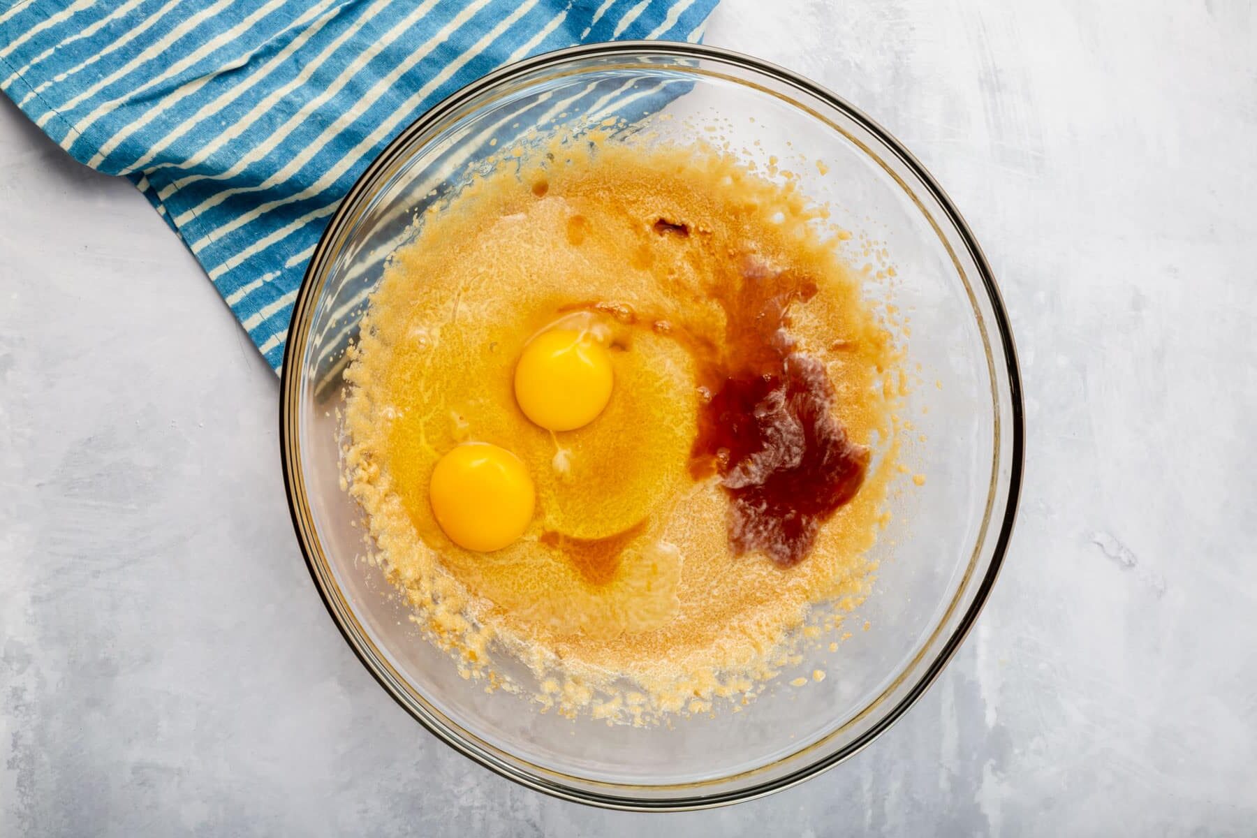 A glass mixing bowl with a creamed butter and sugar mixture topped with vanilla extract and two eggs.