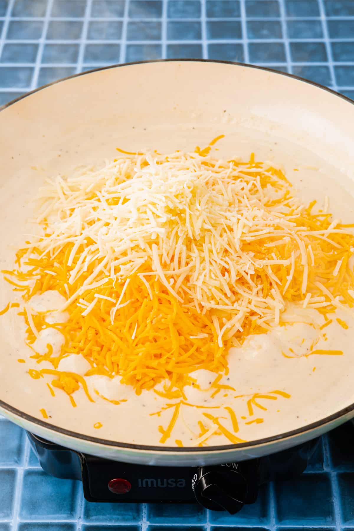 A white braising pan with a white creamy sauce topped with shredded cheddar cheese and parmesan cheese before mixing together.