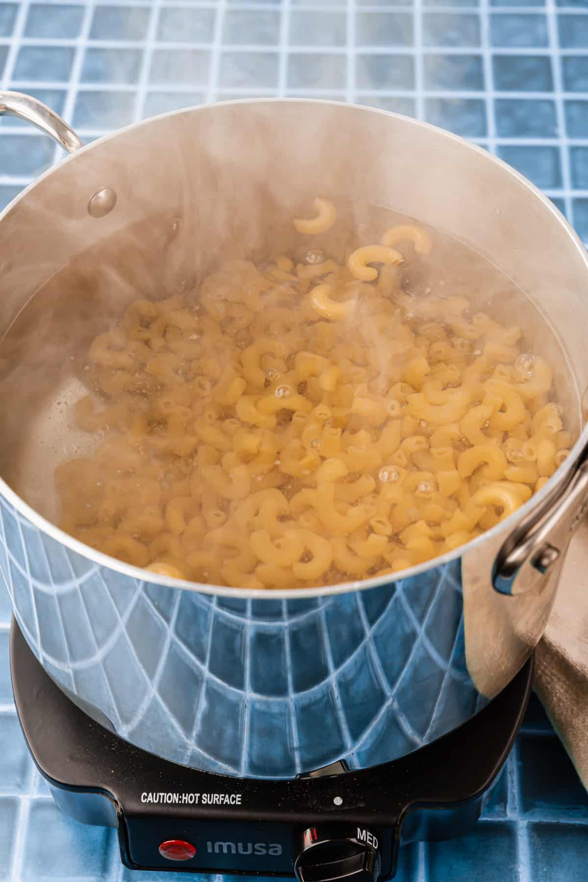 A stainless steel pot of water cooking gluten free macaroni noodles on a hot plate.