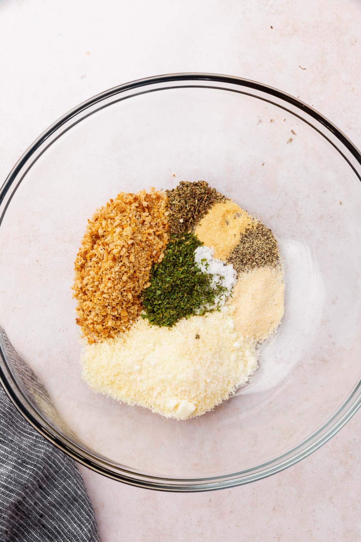 A glass mixing bowl with gluten-free breadcrumbs, grated parmesan, garlic powder, black pepper, dried parsley, dried oregano, onion powder, and salt before mixing together.