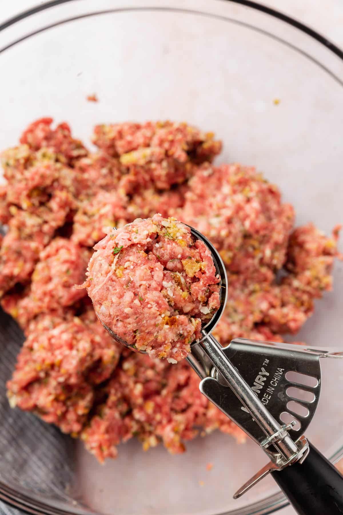 A ice cream scoop with raw gluten-free meatball mixture over a bowl of more meat.