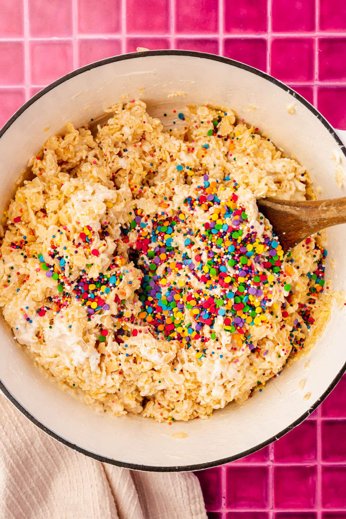 A white dutch oven filled with a gluten free rice krispies treat mixture topped with a pile of rainbow sprinkles.