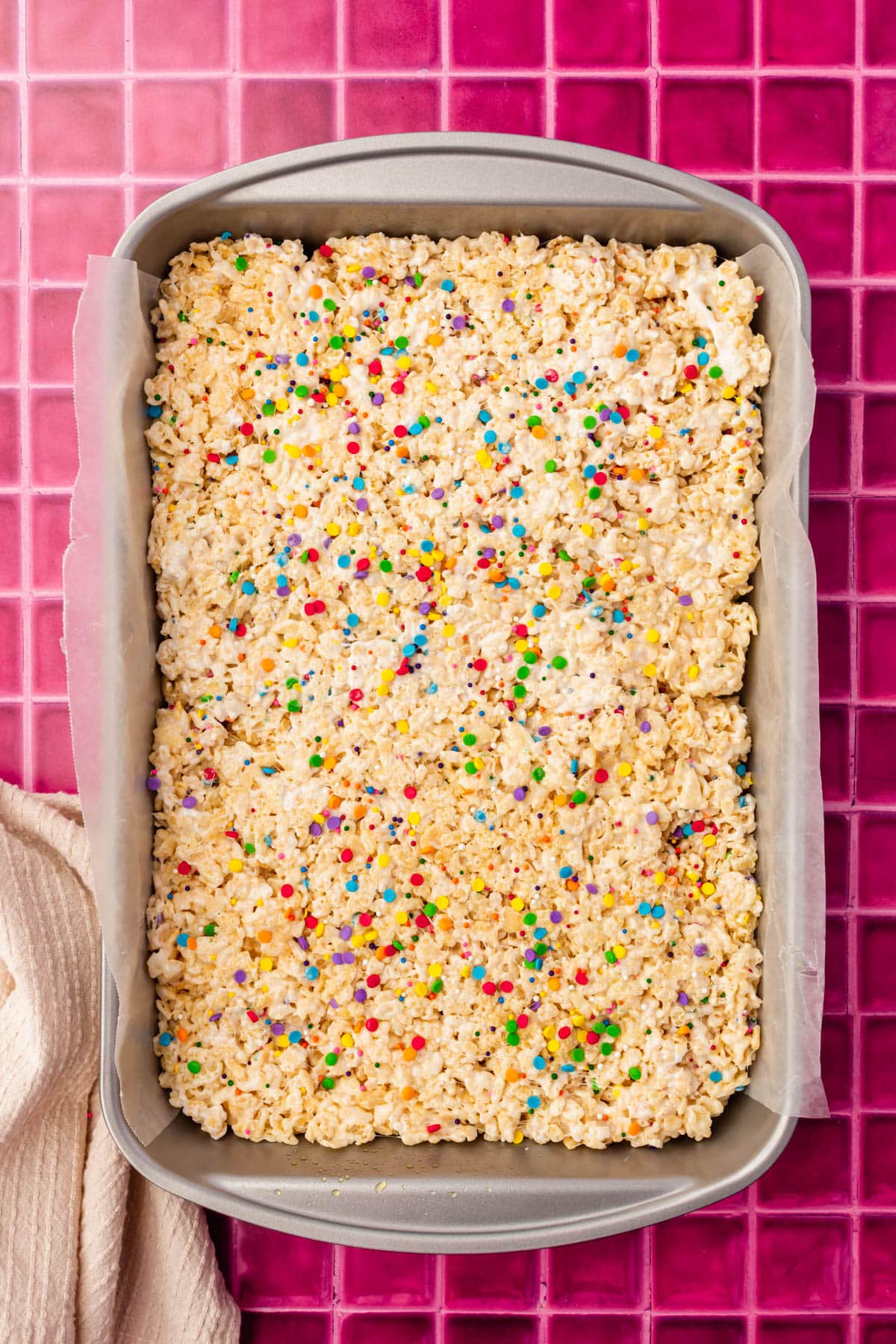 A rectangle metal baking pan lined with wax paper filled with gluten free rice krispies treats toped with rainbow sprinkles.