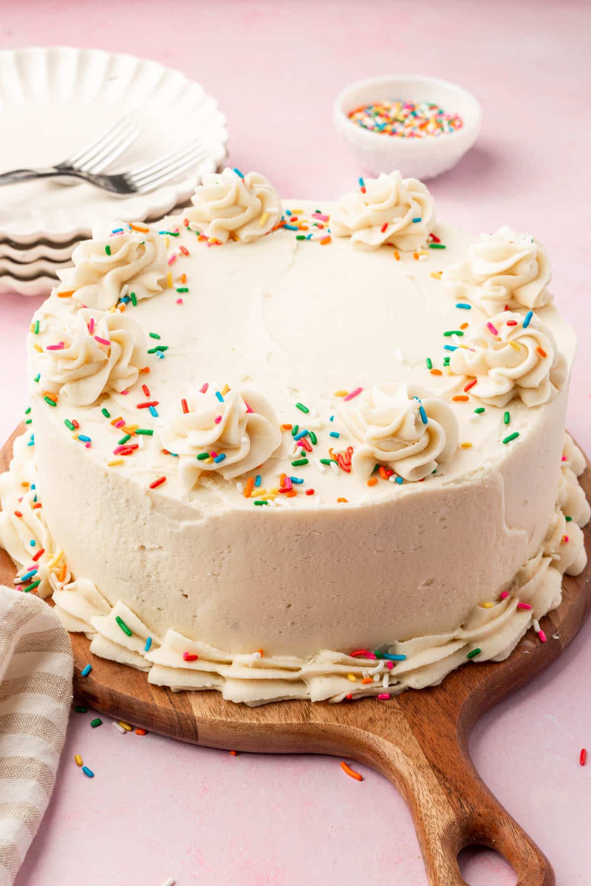A round gluten free vanilla cake with vanilla frosting and rainbow sprinkles on a wood serving board.