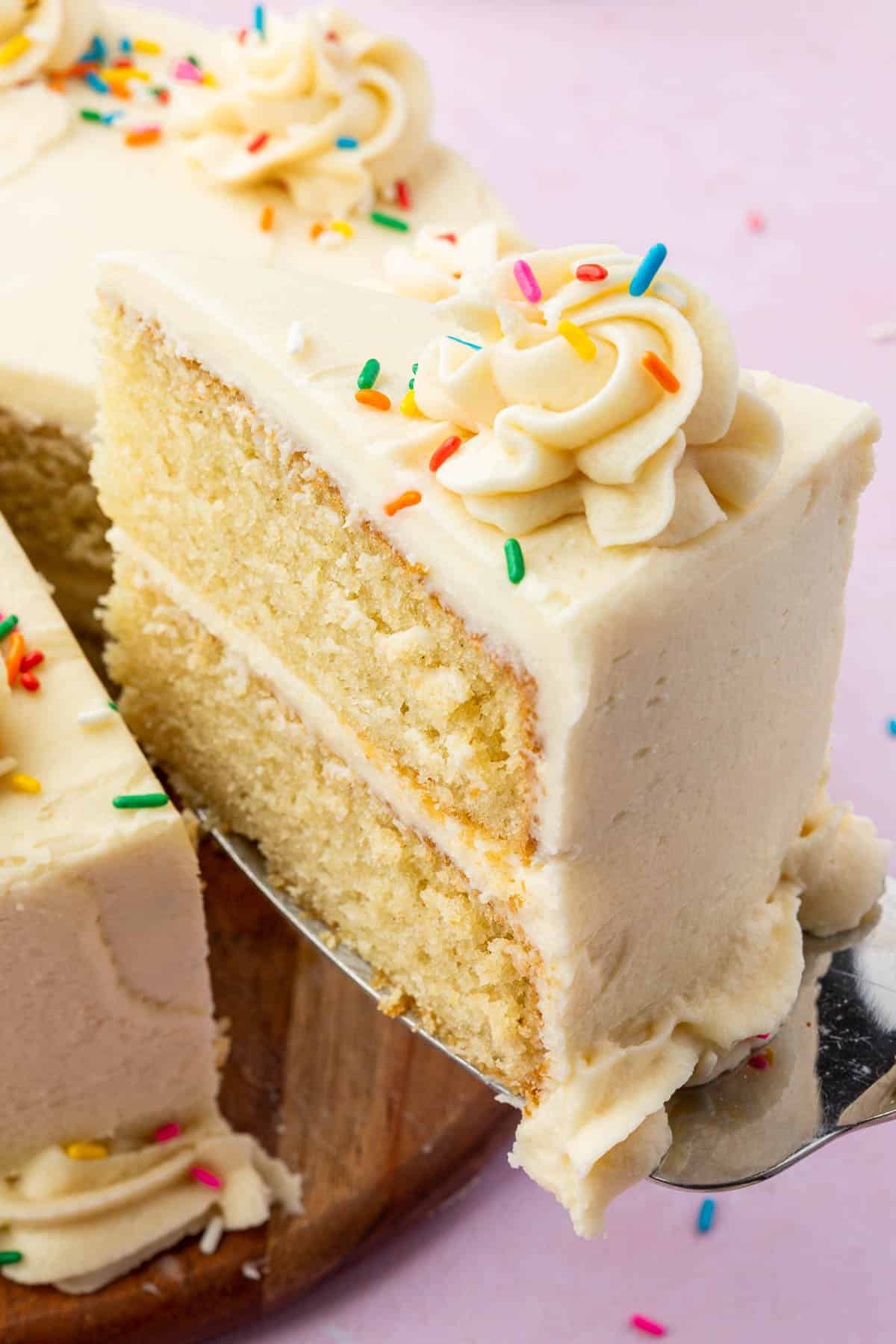 A single slice of 2-layer gluten free vanilla cake topped with rainbow sprinkles being lifted out of the larger cake.