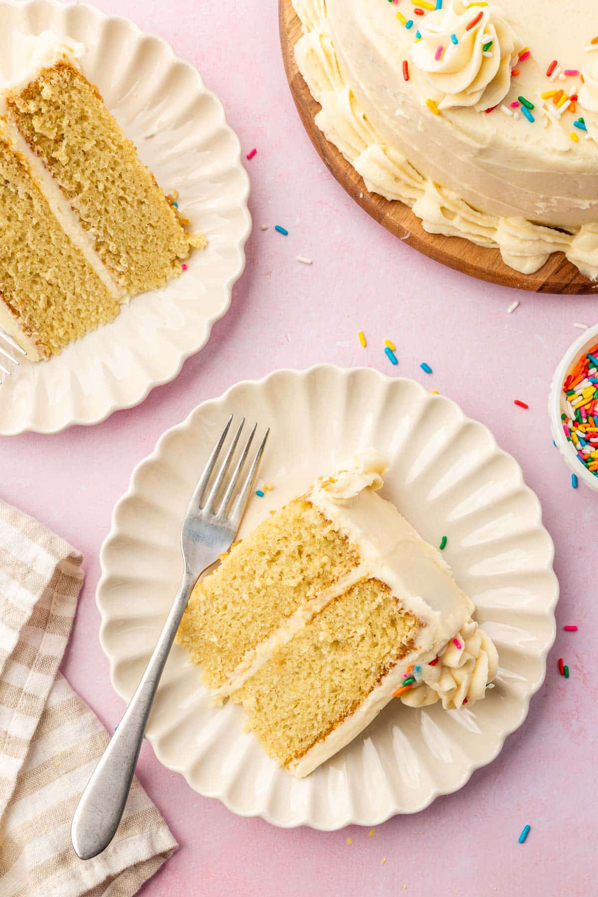 An overhead view of two dessert plates each with a single slice of gluten free vanilla cake with the larger cake peaking in to the photo.