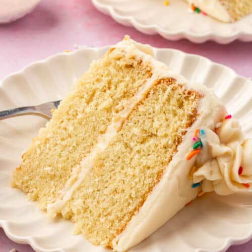 A slice of 2-layer gf vanilla cake with vanilla frosting and rainbow sprinkles on a dessert plate with another slice of cake and a bowl of rainbow sprinkles in the background.