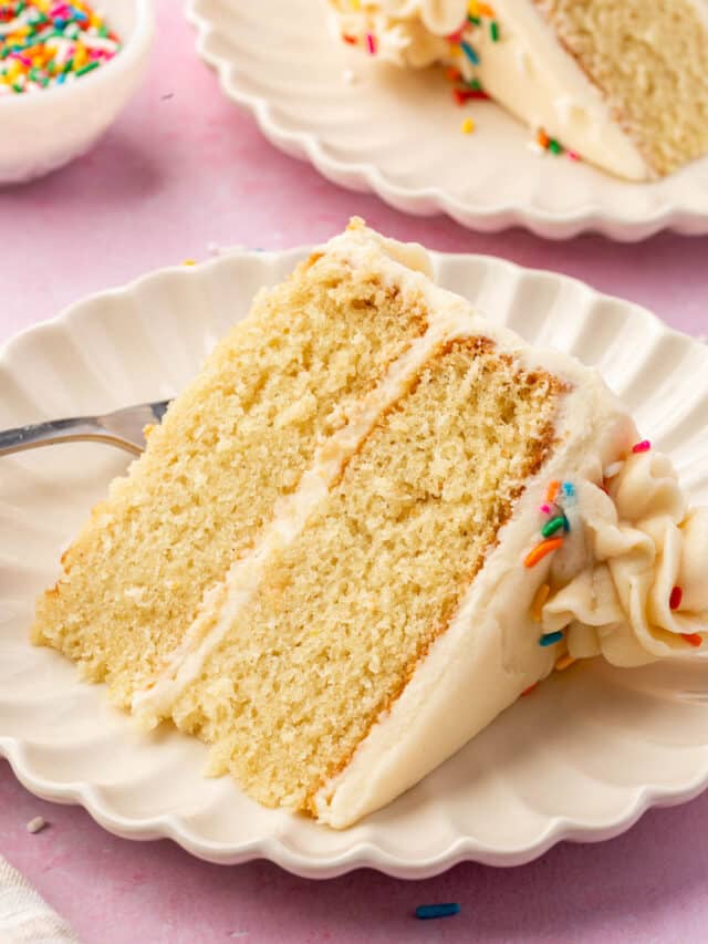 A slice of 2-layer gf vanilla cake with vanilla frosting and rainbow sprinkles on a dessert plate with another slice of cake and a bowl of rainbow sprinkles in the background.