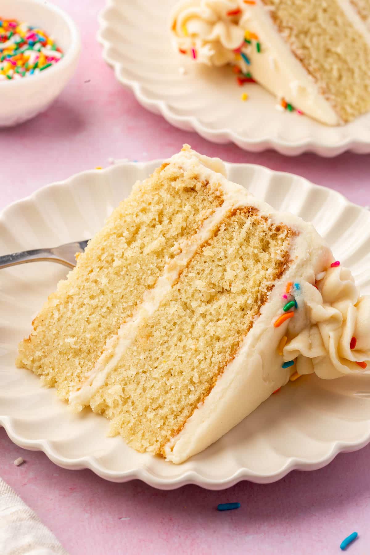 A slice of 2-layer gf vanilla cake with vanilla frosting and rainbow sprinkles on a dessert plate with another slice of cake and a bowl of rainbow sprinkles in the background.