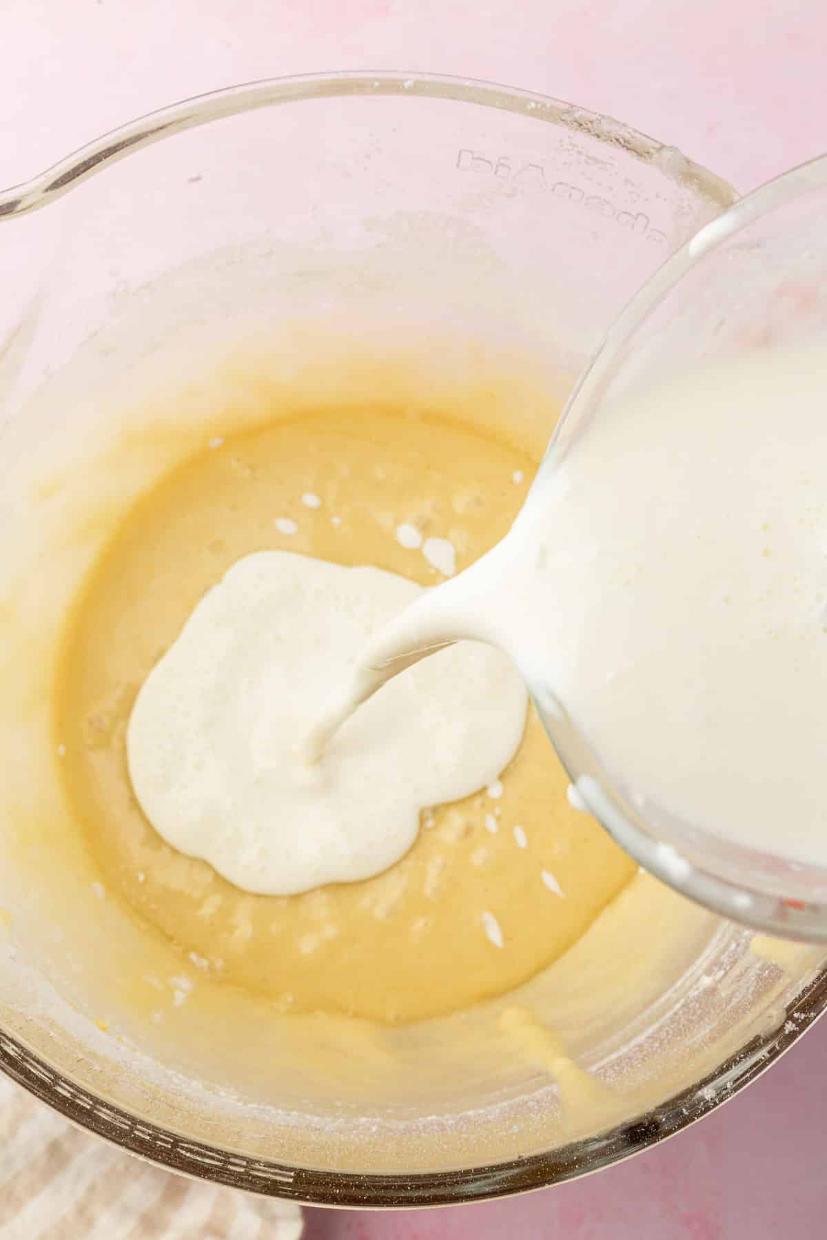 A glass measuring cup pouring a sour cream and milk mixture into a mixing bowl of gluten free cake batter.