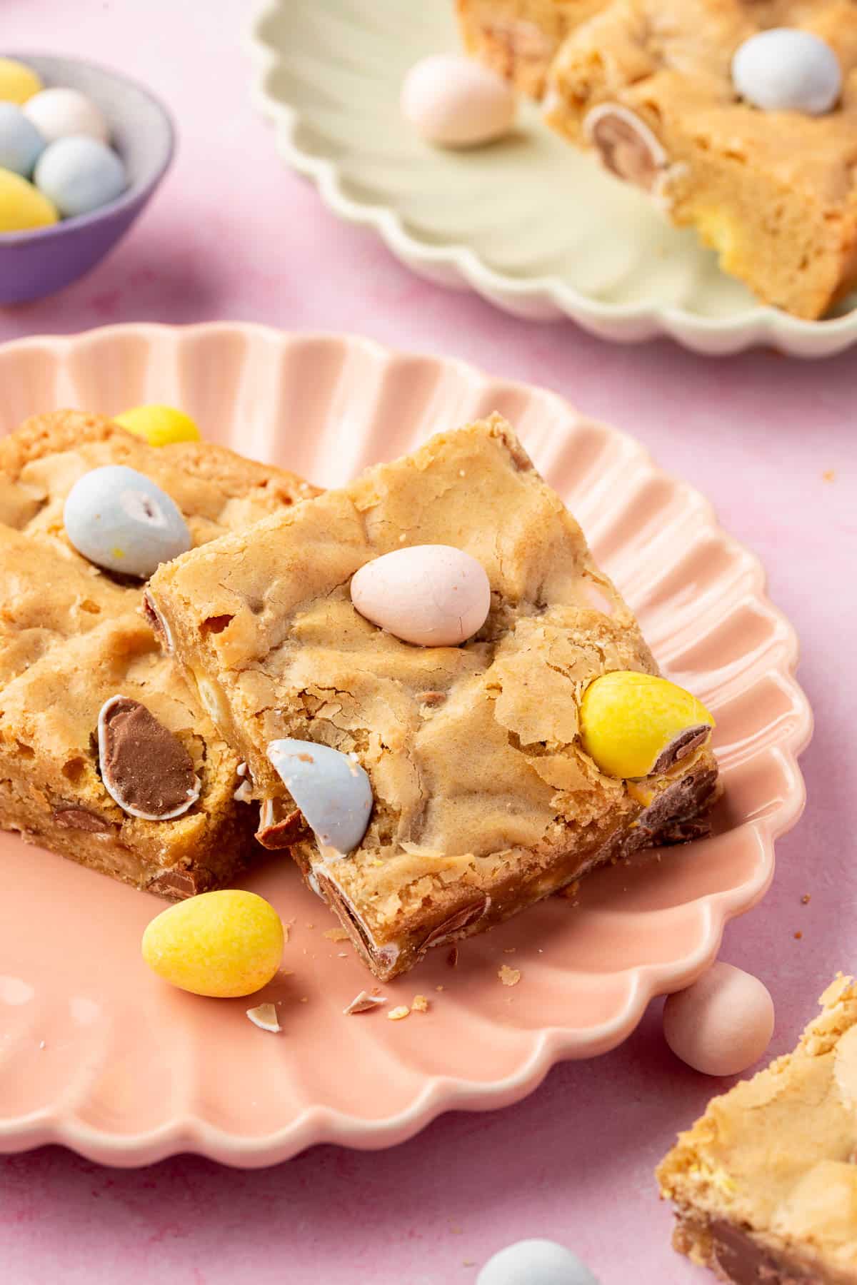 Two gluten-free mini egg blondies leaning against each other on a pink scalloped dessert plate with additional blondies and mini chocolate eggs in the background.