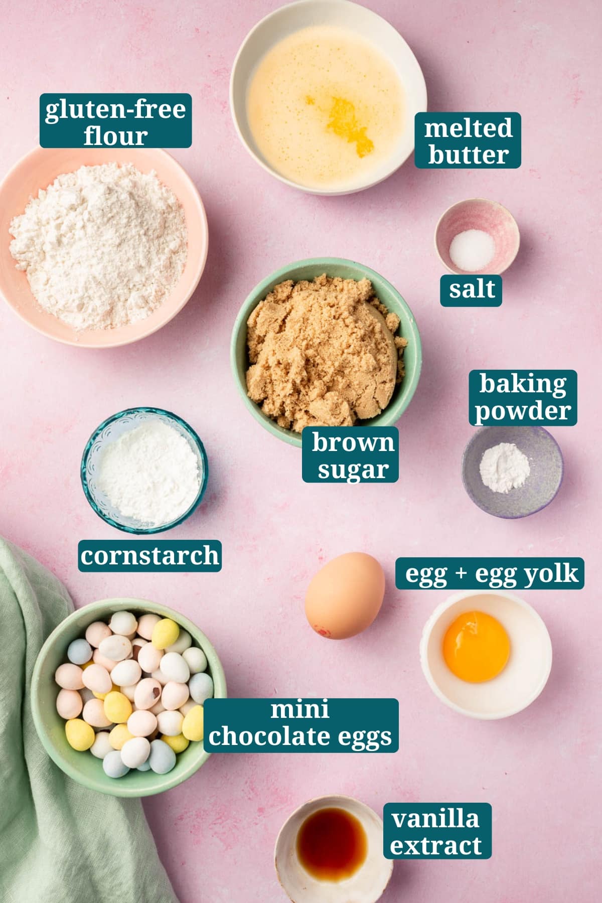 An overhead view of ingredients in small bowls to make gluten-free mini egg blondies, including melted butter, gluten-free flour, brown sugar, cornstarch, baking powder, salt, egg, egg yolk, mini chocolate eggs, and vanilla extract with text overlays over each ingredient to say what it is.