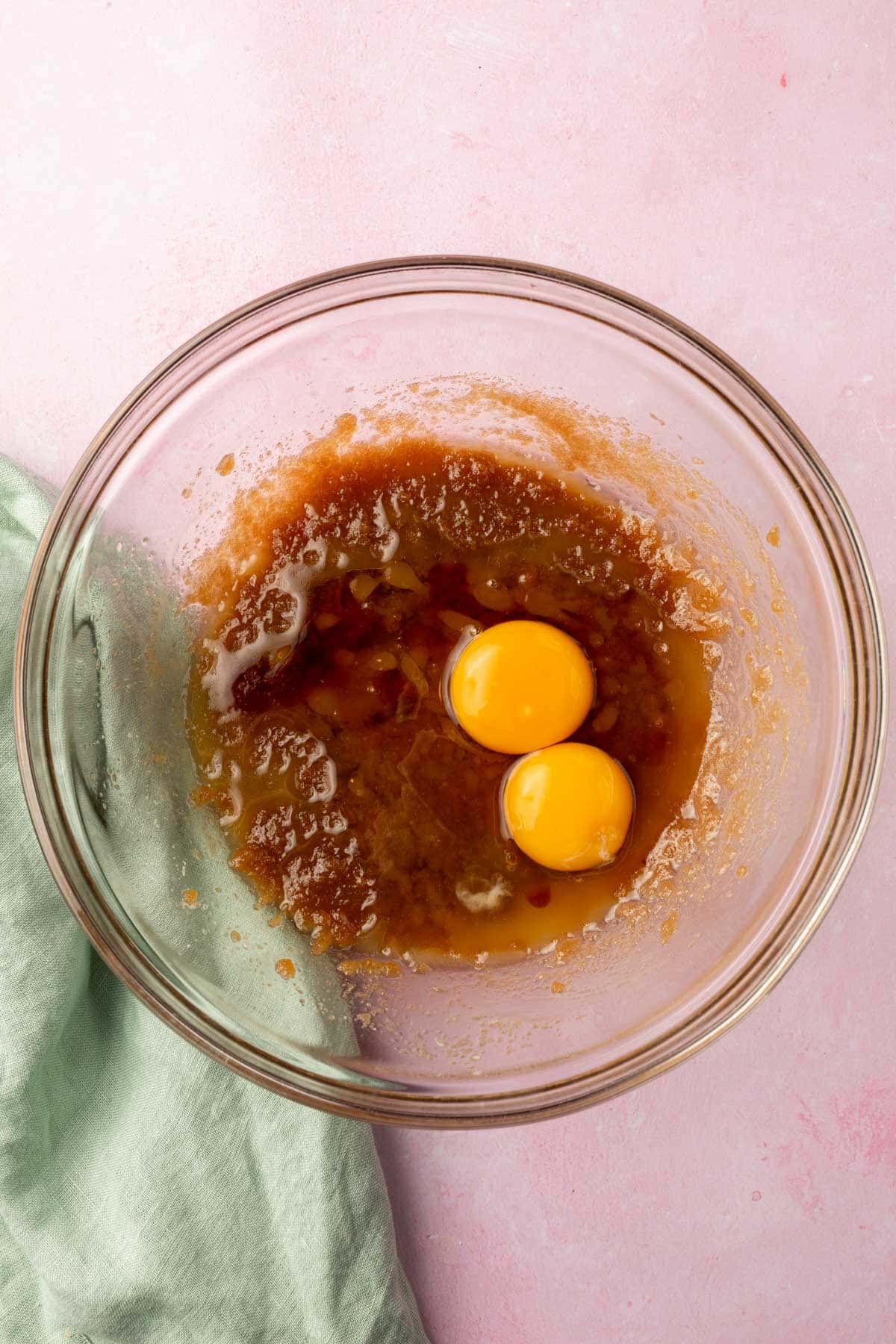 A glass mixing bowl with a brown sugar and melted butter mixture topped with two eggs and vanilla extract before mixing together.