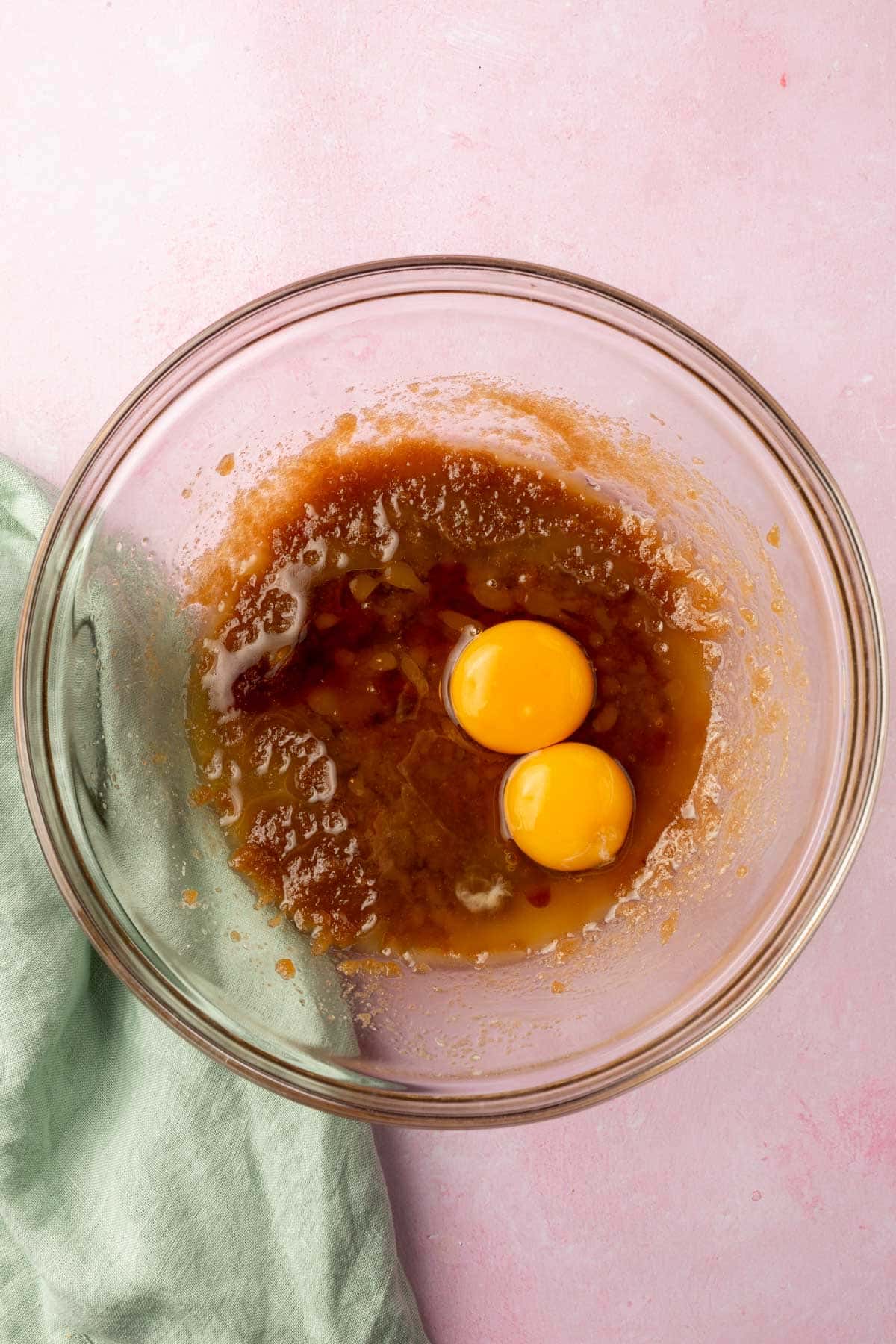 A glass mixing bowl with a brown sugar and melted butter mixture topped with two eggs and vanilla extract before mixing together.