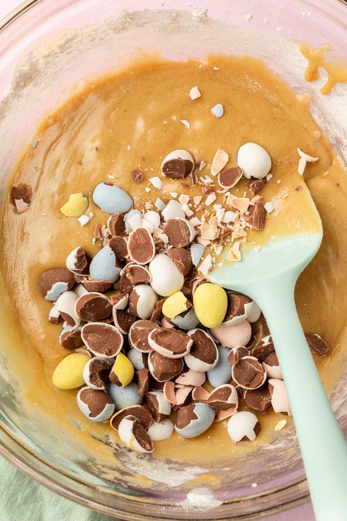 A glass mixing bowl of gluten-free blondie batter topped with a pile of chopped up mini chocolate eggs before mixing together.