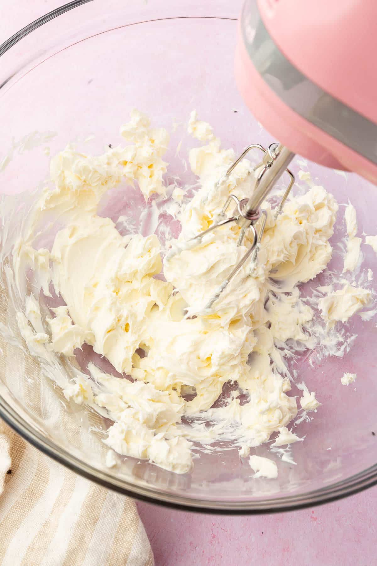 An electric hand mixer blending cream cheese in a glass mixing bowl.