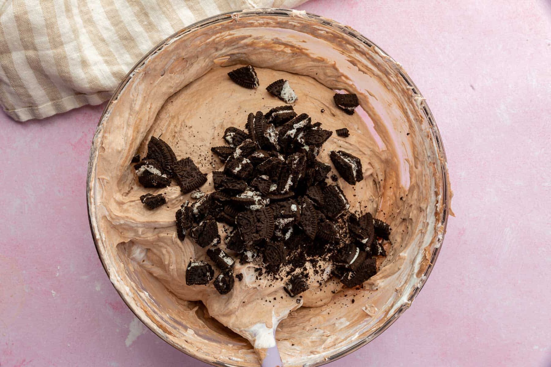A glass mixing bowl with a chocolate whipped pudding mixture topped with a pile of crushed gluten-free Oreos.