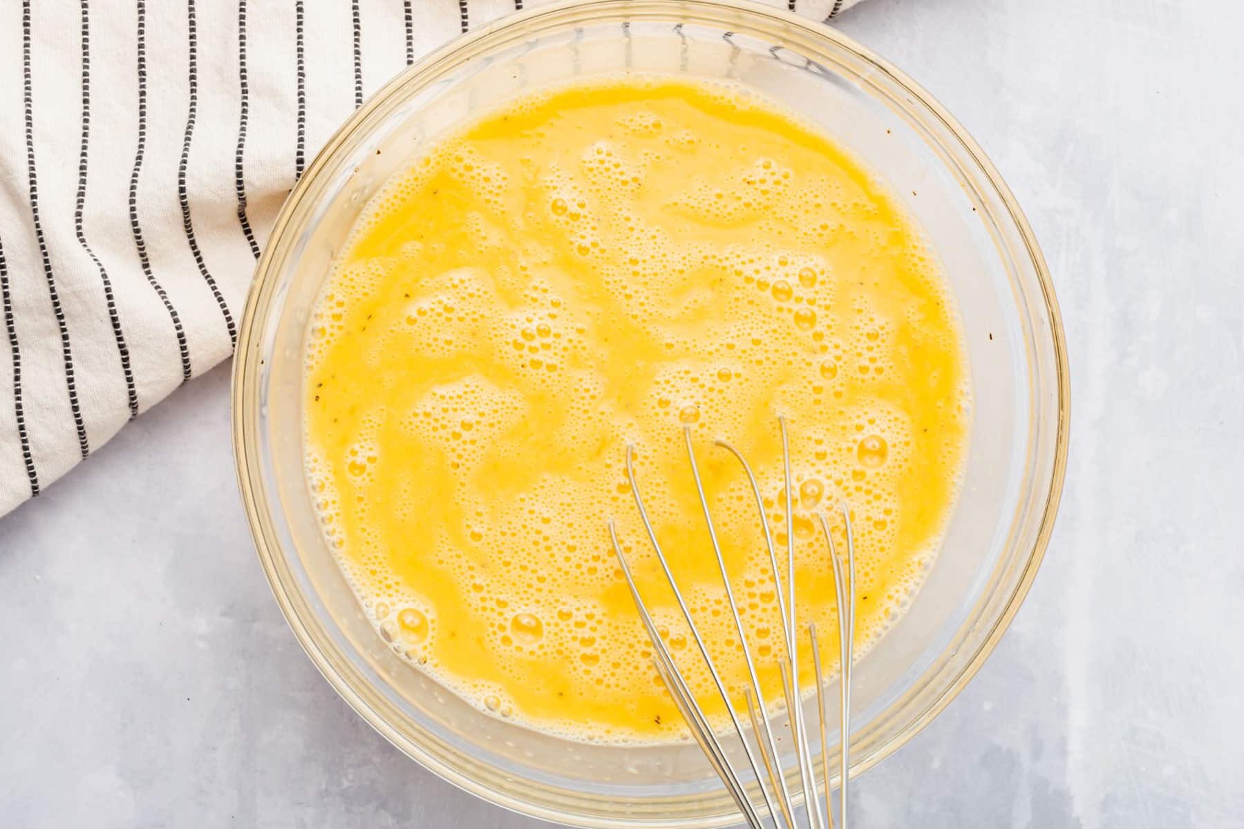 A glass mixing bowl of whisked eggs with a silver whisk.