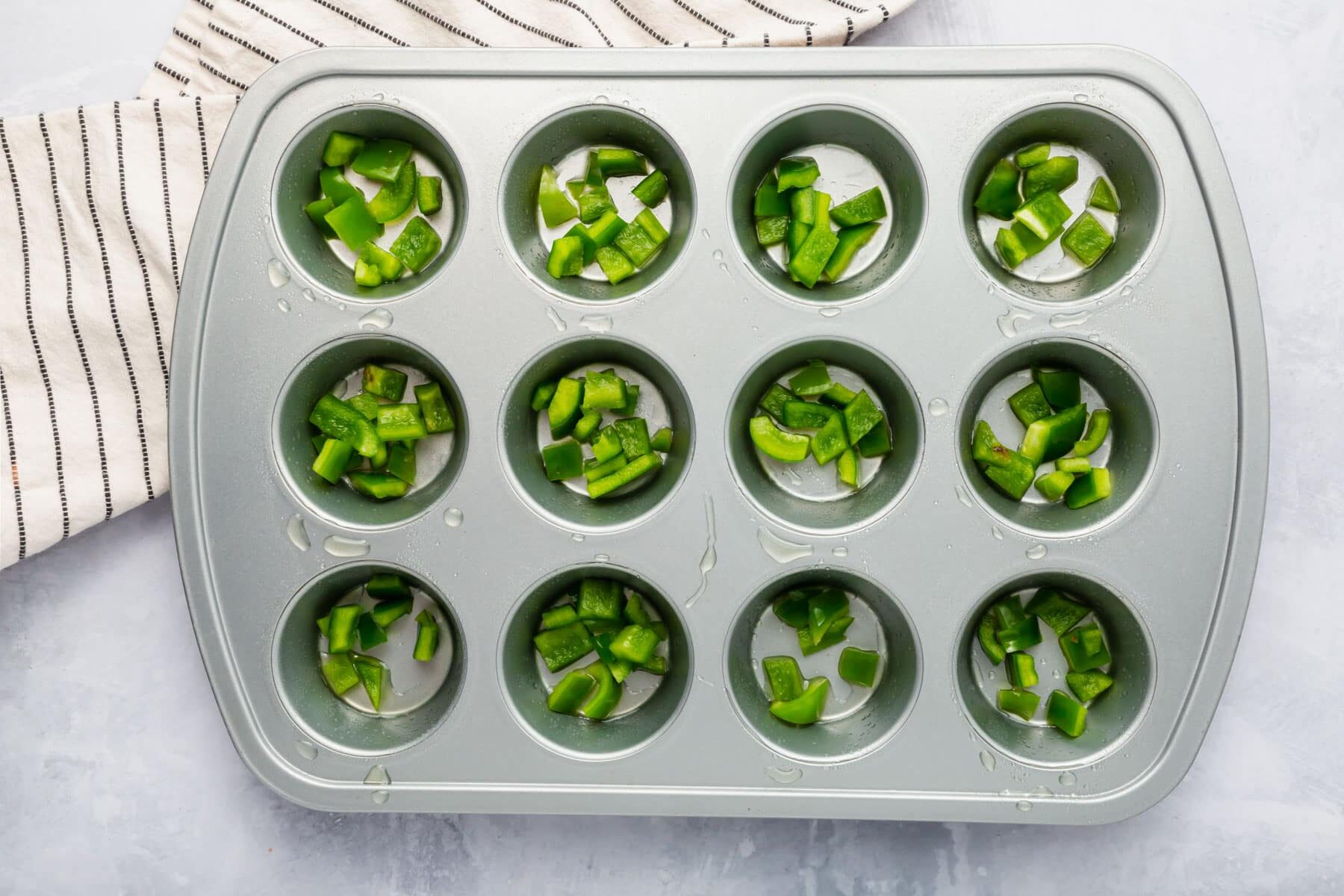 A silver muffin tin filled with chopped green bell pepper in each muffin well.