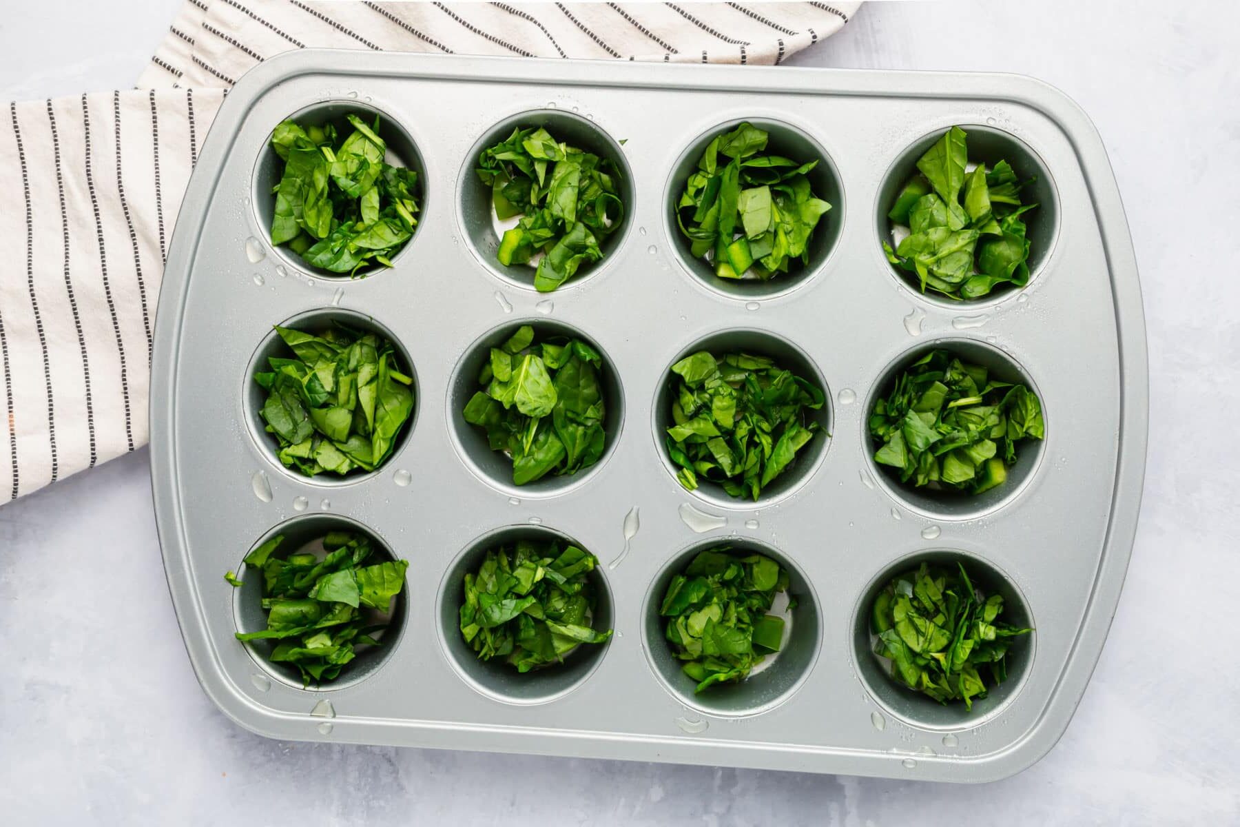 A silver muffin tin filled with roughly chopped spinach in each muffin well.