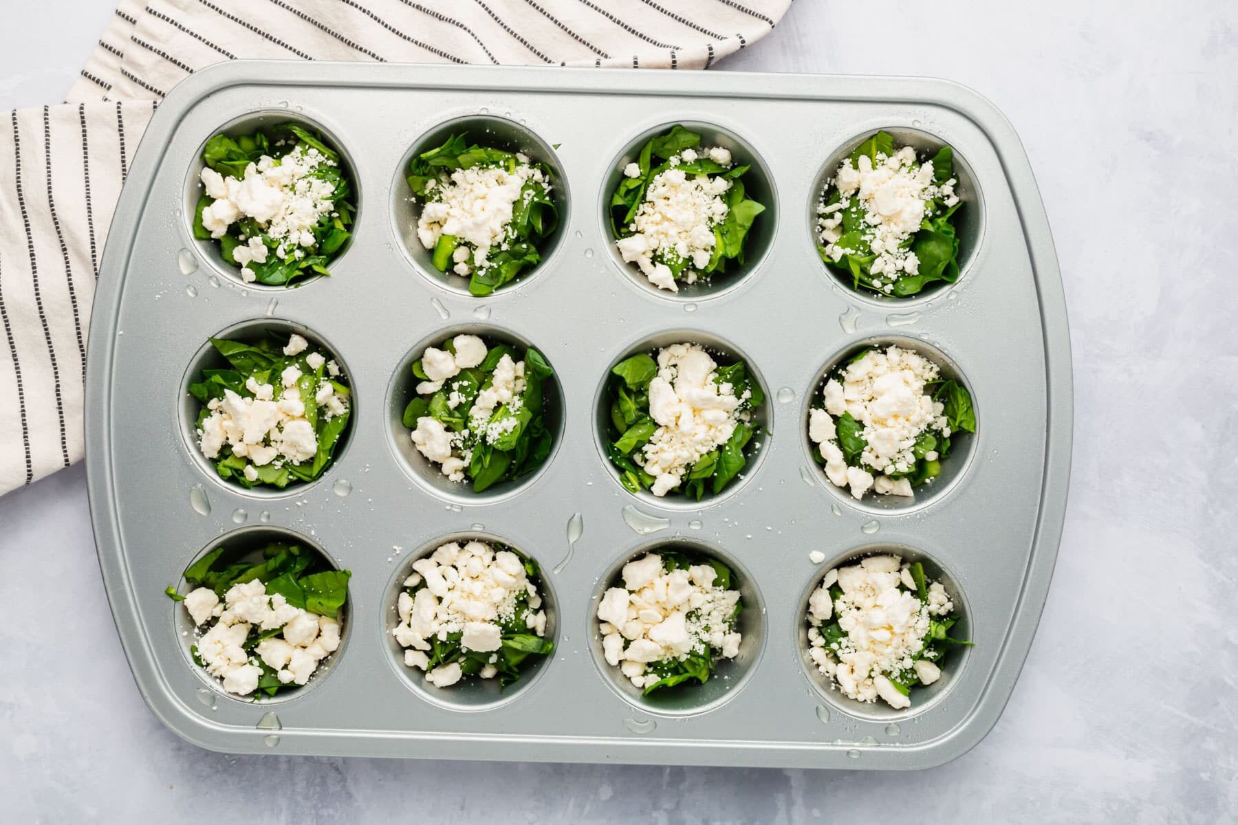 A muffin tin filled with spinach and crumbled feta in each well.