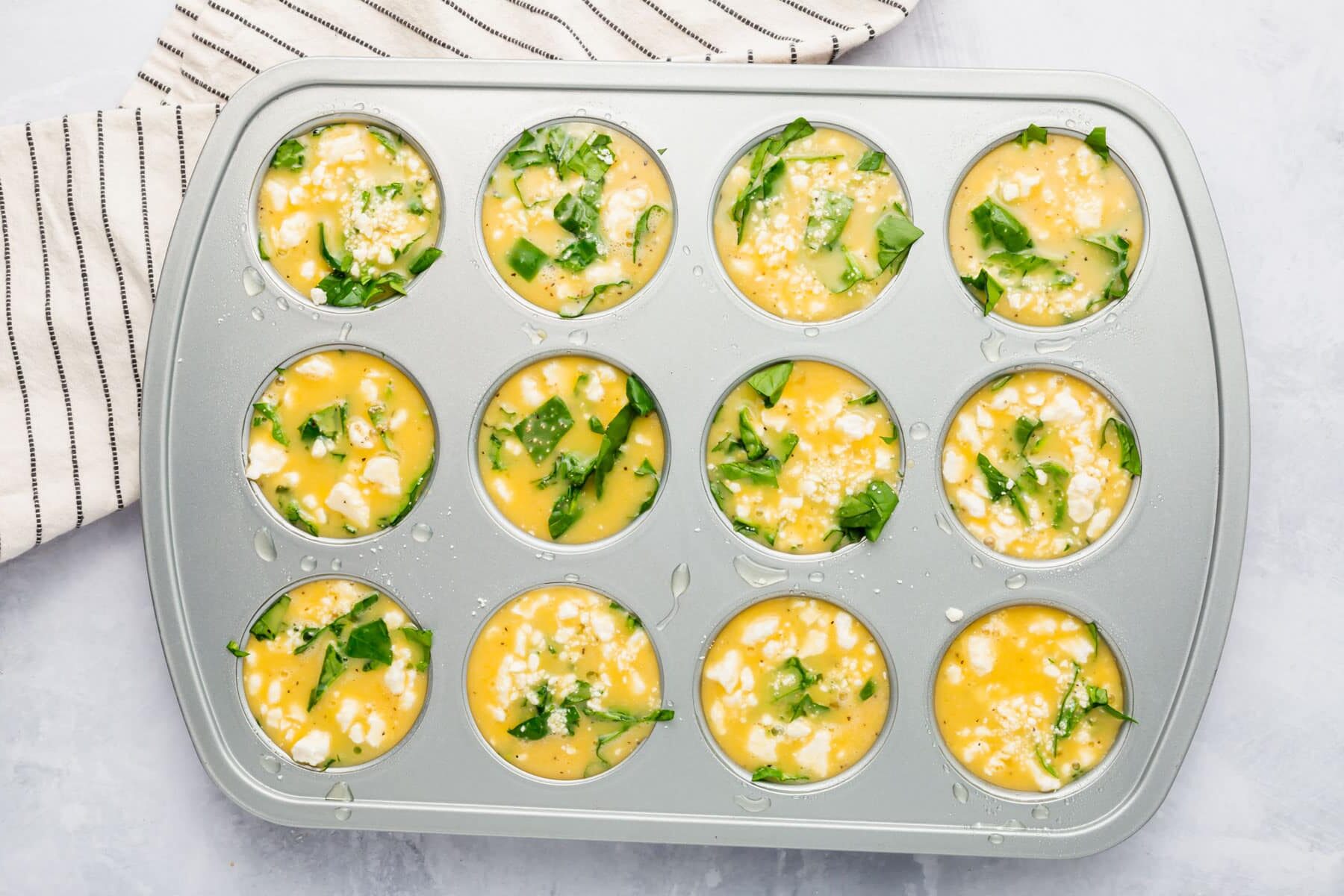 A muffin tin filled with spinach, feta, and an egg mixture before baking in the oven.