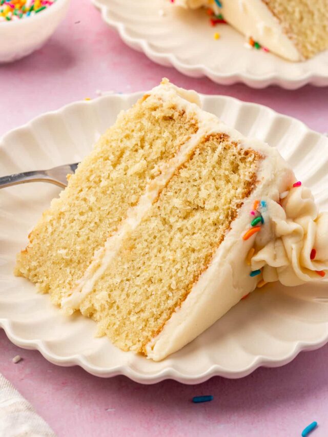 A slice of 2-layer gf vanilla cake with vanilla frosting and rainbow sprinkles on a dessert plate with another slice of cake and a bowl of rainbow sprinkles in the background.