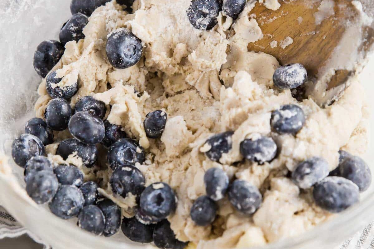 A clear glass bowl filled with a thick, partially mixed dough and fresh blueberries, being stirred with a wooden spoon on a gray and white striped cloth.