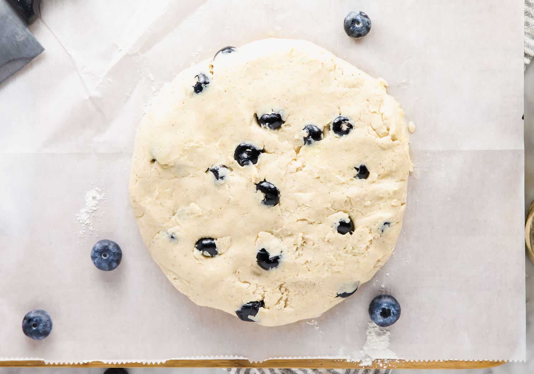 A round disk of unbaked dough with blueberries scattered throughout, resting on a sheet of parchment paper. A few fresh blueberries are placed around the dough.
