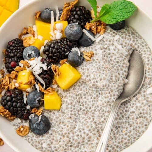 A chia pudding bowl with blackberries, blueberries, mango, coconut, and granola with a spoon in the bowl.