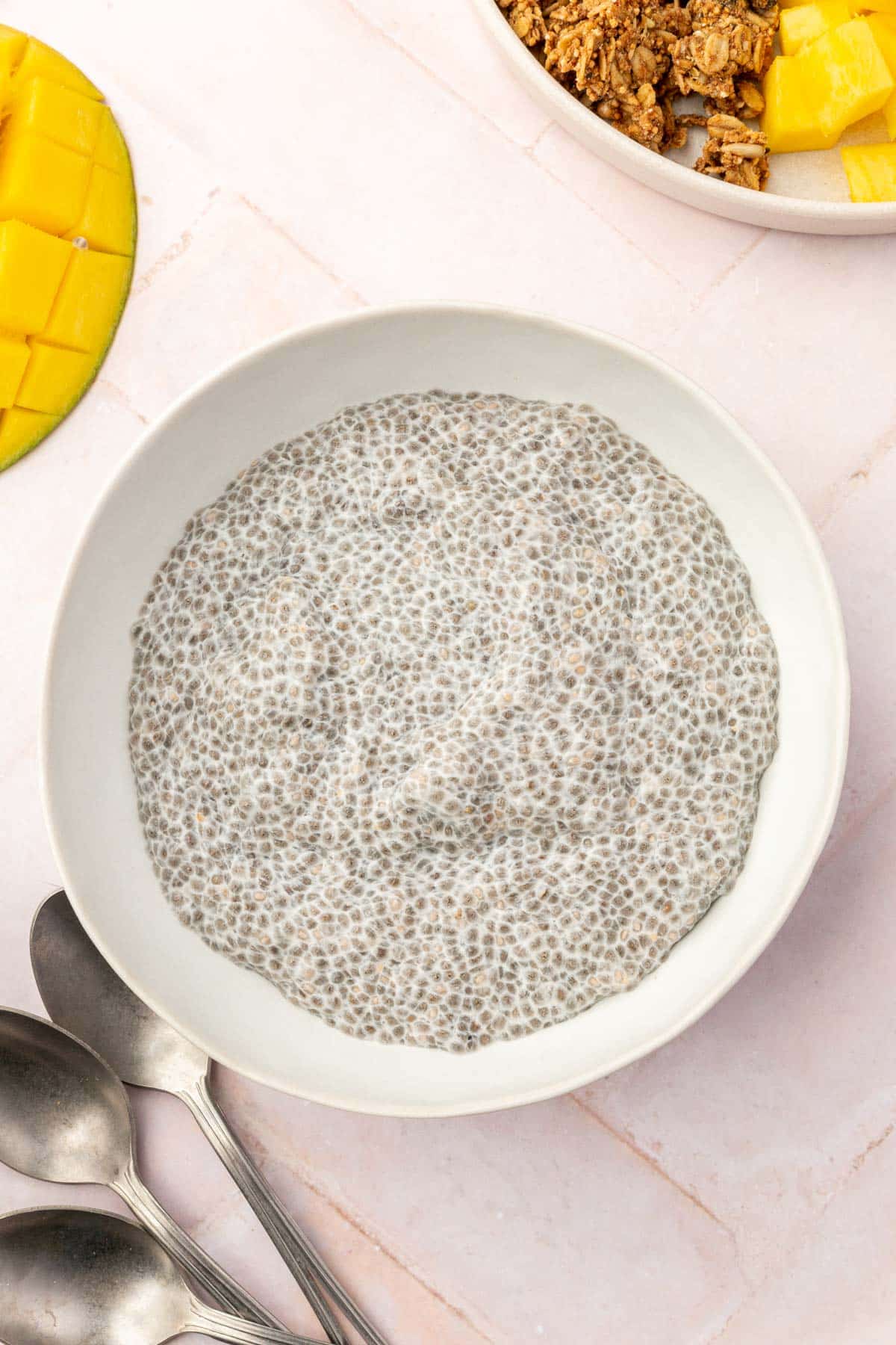 A bowl of chia pudding with spoons to the side and a diced mango to the side.