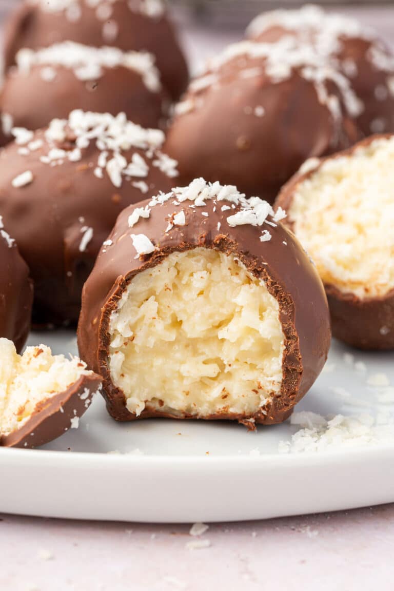 A chocolate covered coconut ball that has a bite taken out of it on a white plate with more truffles in the backbround.