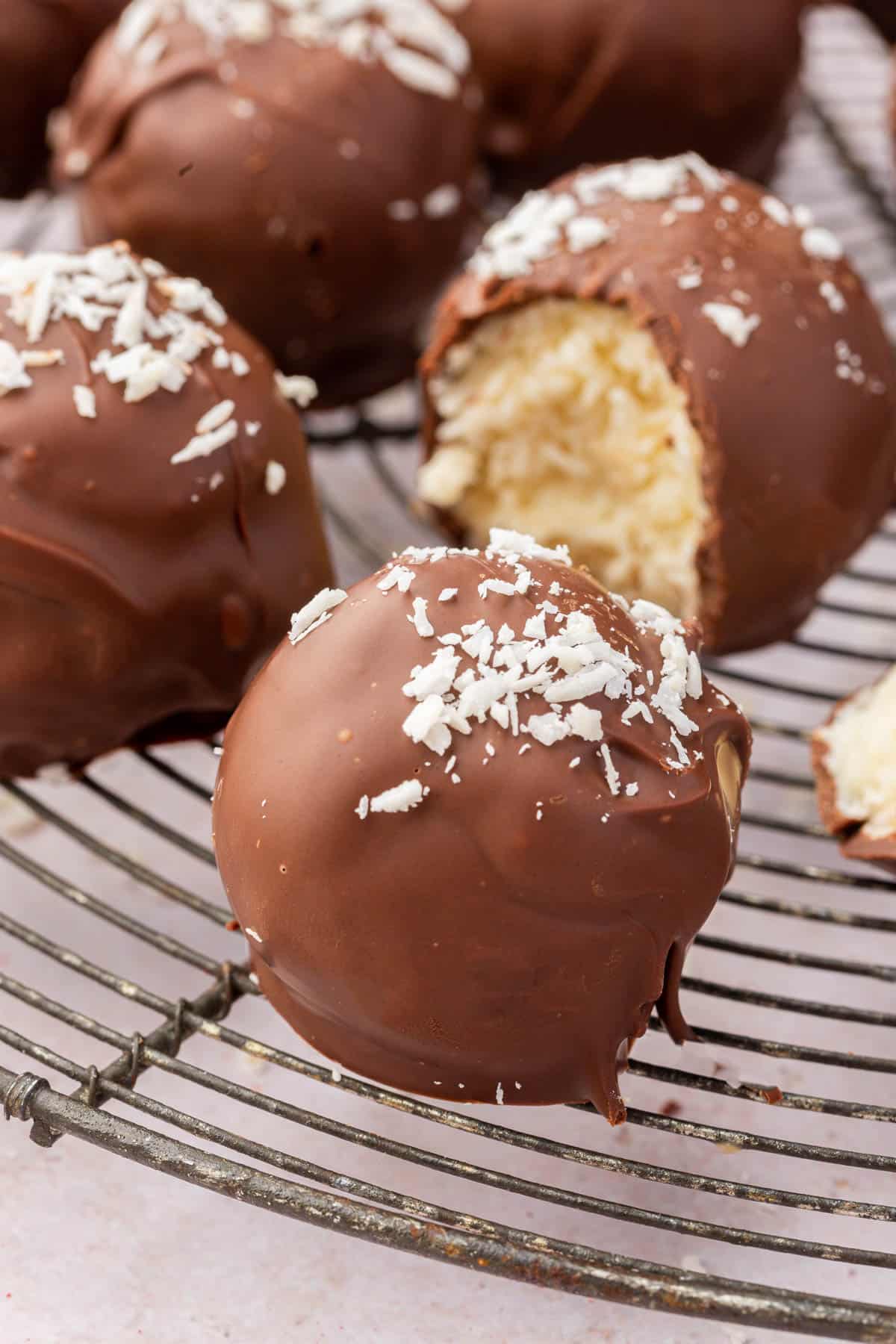 A close up of a single coconut ball covered in dark chocolate and topped with shredded coconut flakes on a cooling rack with more coconut balls in the background.
