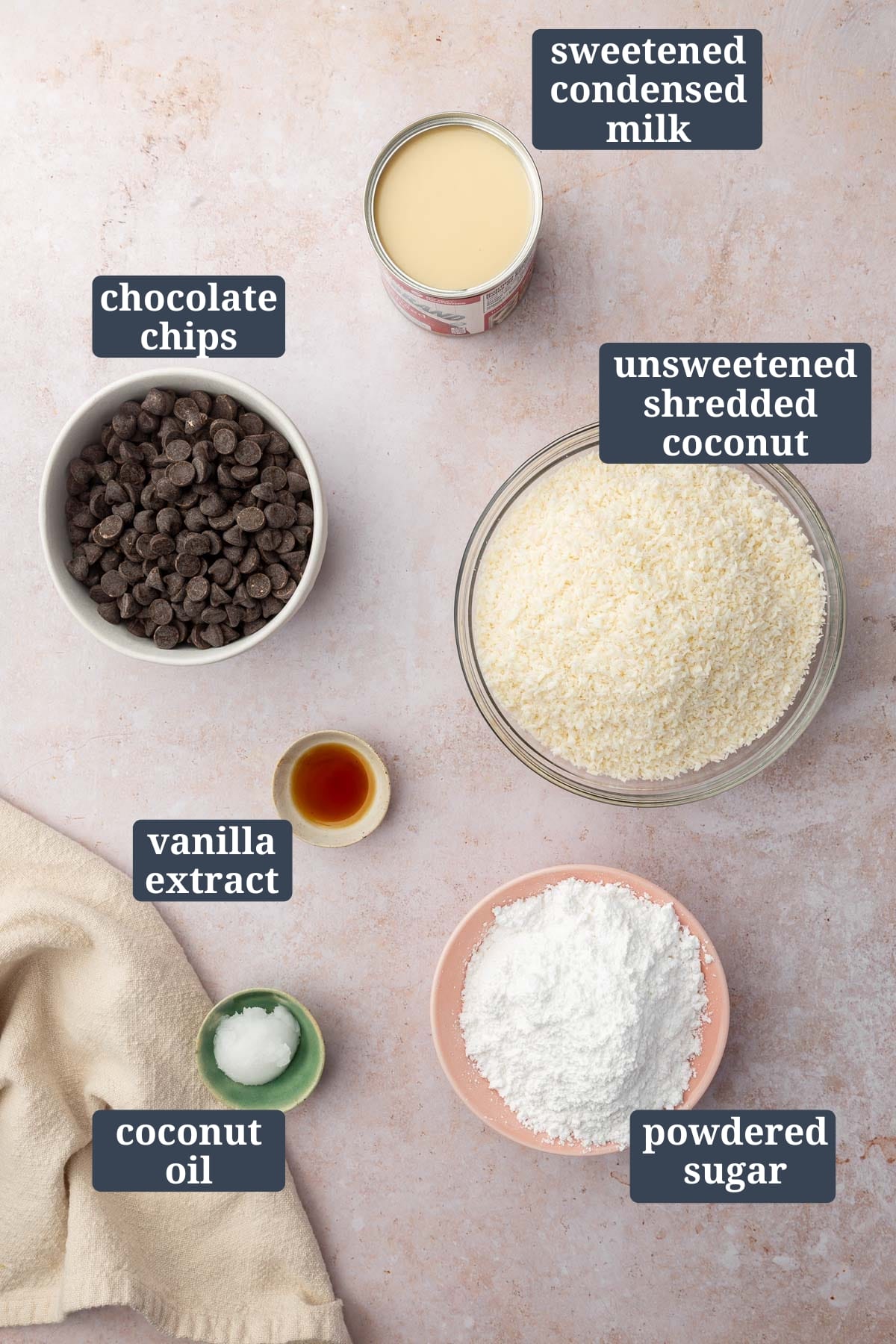 An overhead view of ingredients to make coconut balls including sweetened condensed milk, chocolate chips, unsweetened shredded coconut, vanilla extract, coconut oil, and powdered sugar with text overlays over each ingredient to say what it is.