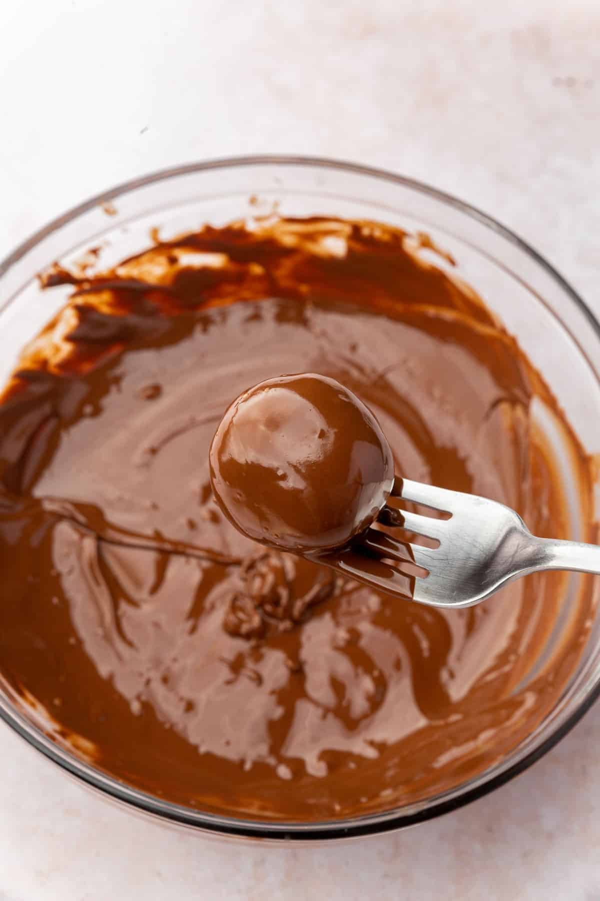 A coconut ball that has been dipped into a bowl of melted dark chocolate and pulled up with a fork.