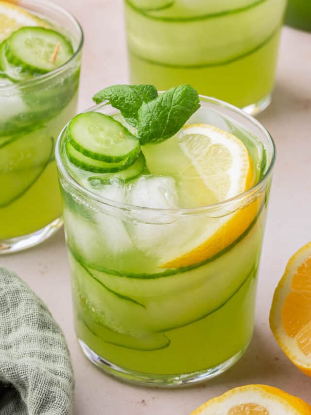 A glass of cucumber lemonade with ice, a lemon wedge, cucumber ribbons and slices and a sprig of mint in it.