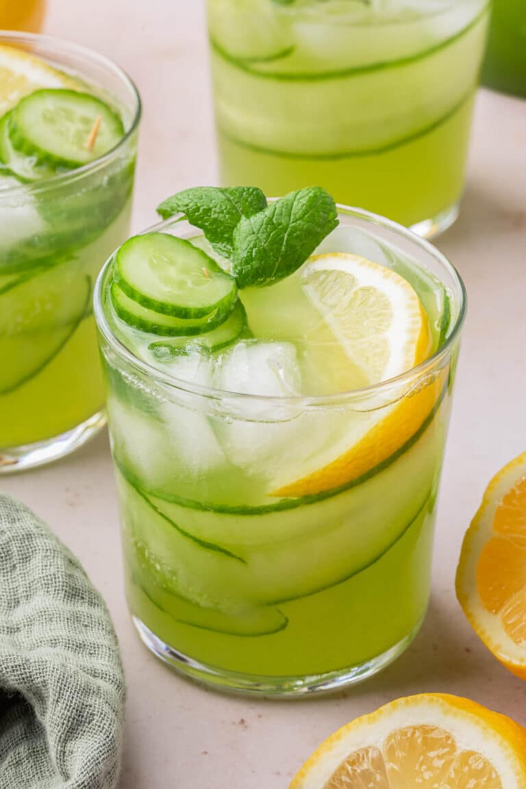 A glass of cucumber lemonade with ice, a lemon wedge, cucumber ribbons and slices and a sprig of mint in it.