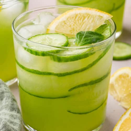 A glass of cucumber lemonade with ice, a lemon wedge and slices of cucumber with more glasses in the background.