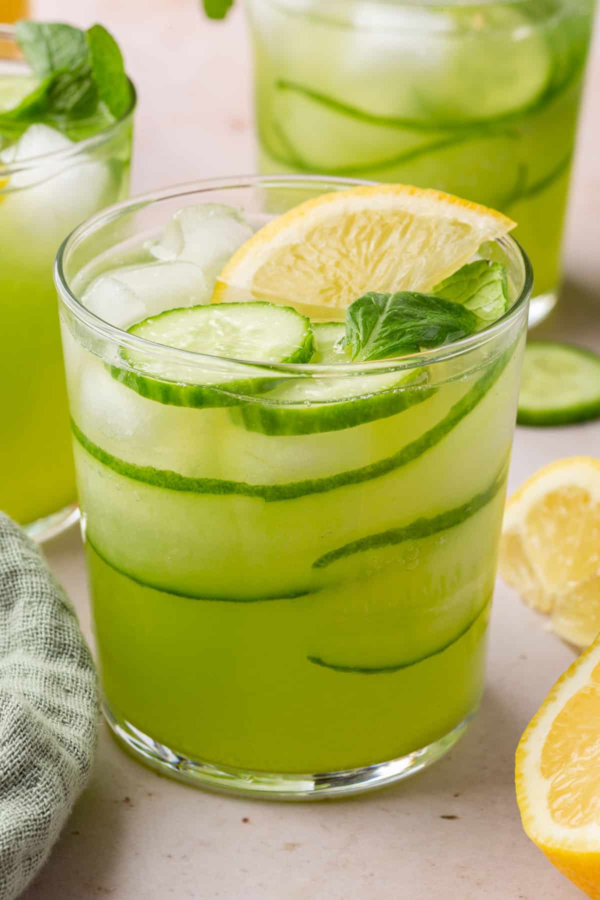 A glass of cucumber lemonade with ice, a lemon wedge and slices of cucumber with more glasses in the background.