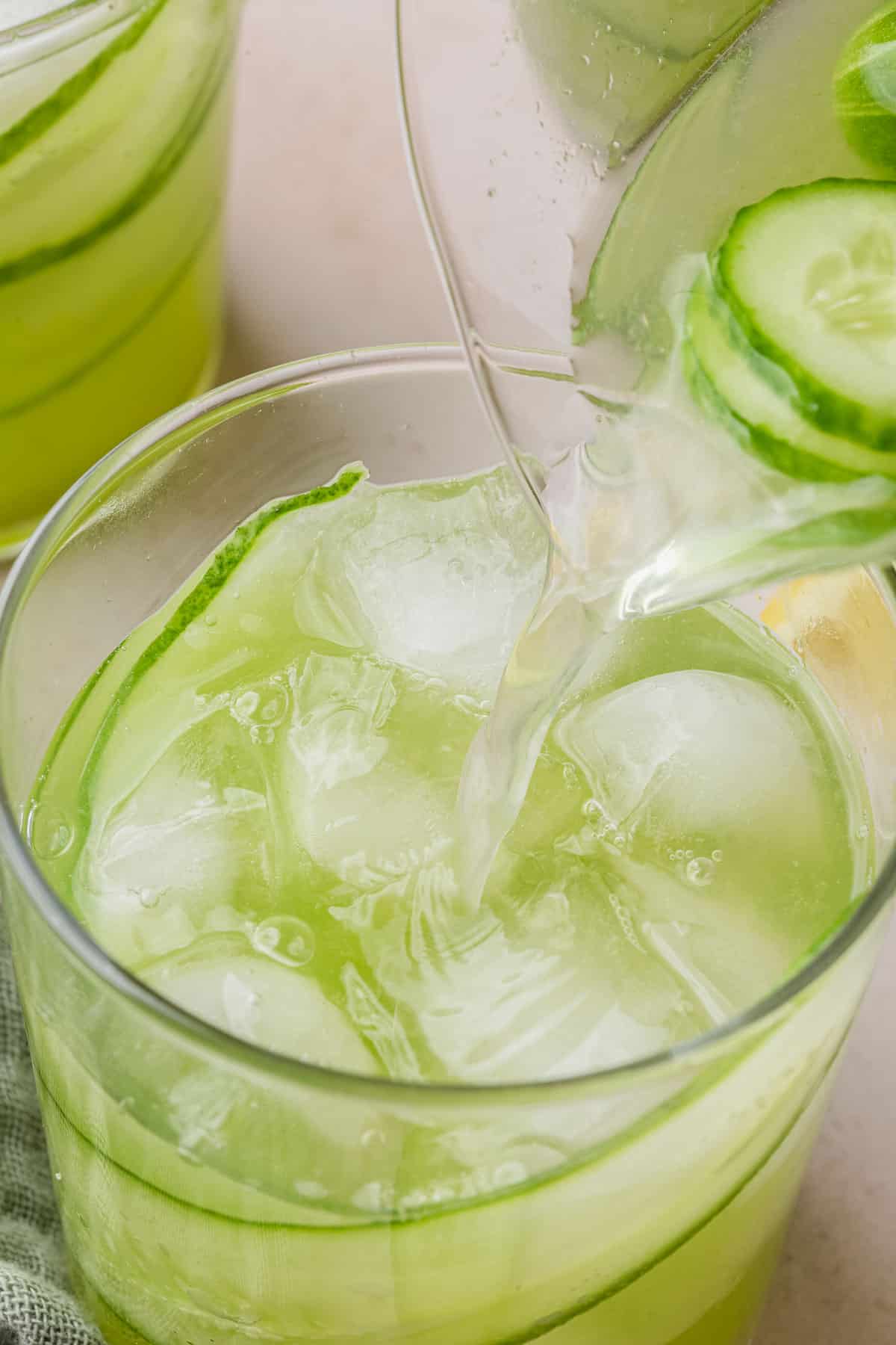 A pitcher pouring cucumber lemonade into a glass with ice.